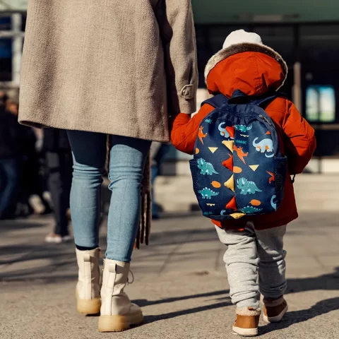 Rückansicht eines kleinen Kindes mit Dinosaurier-Rucksack an der Hand eines Erwachsenen, die gemeinsam auf einem sonnigen Platz gehen.