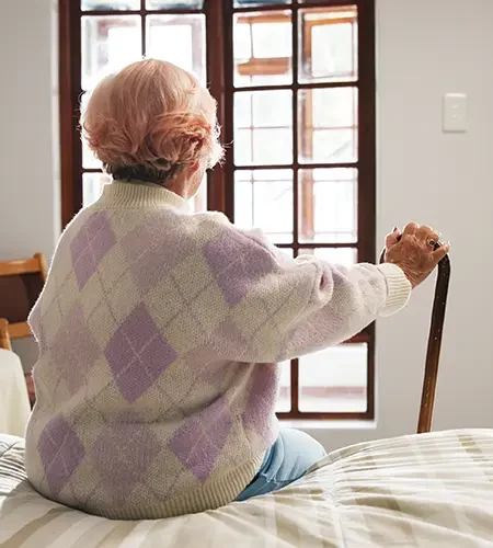 Rückansicht einer älteren Frau mit rosa getöntem Haar, die in einem hellen Zimmer auf einer Bettkante sitzt und sich mit einer Hand auf einen Gehstock stützt, während sie aus dem Fenster blickt.
