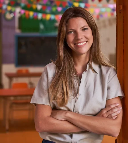 Eine freundlich lächelnde Frau mit langen braunen Haaren steht mit verschränkten Armen in einem Kindergartenraum. Im unscharfen Hintergrund sind Tische, Stühle und eine bunte Wimpelkette zu erkennen.