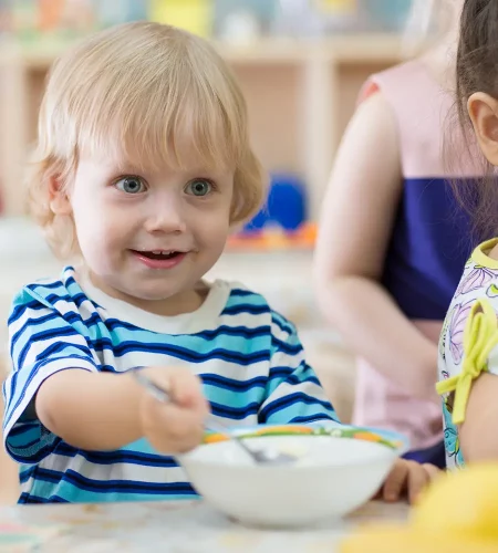 Nahaufnahme eines kleinen Kindes mit hellen Haaren und blauen Augen, das mit einem Löffel in einer Müslischale rührt. Das Kind blickt lächelnd zur Seite, im Hintergrund sind vage andere Kinder zu sehen.