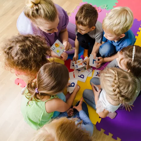 Vogelperspektive auf eine Erzieherin und sechs Kinder, die gemeinsam auf einem farbenfrohen Puzzle-Teppich sitzen. Sie schauen konzentriert auf Bild- und Buchstabenkarten, die in der Mitte verteilt liegen.