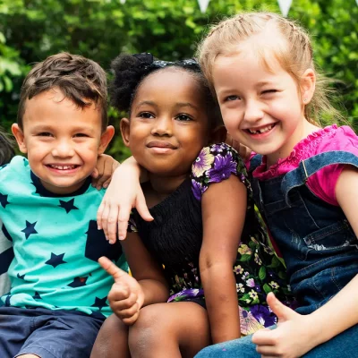 Drei Kinder unterschiedlicher Herkunft sitzen Arm in Arm vor grünem Gebüsch. Sie strahlen in die Kamera und zeigen teilweise den ‚Daumen hoch‘-Geste als Zeichen von Freundschaft und Spaß.