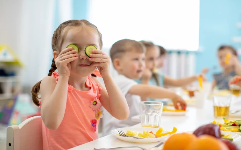 Ein junges Mädchen mit geflochtenen Zöpfen sitzt fröhlich an einem gedeckten Esstisch und hält sich zwei Gurkenscheiben wie eine Brille vor die Augen. Im Hintergrund sitzen weitere Kinder an der Tafel und essen gemeinsam.