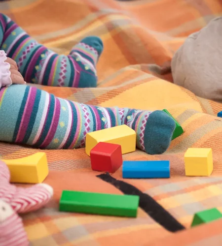 Detailansicht eines Babys auf einer orange-karierten Decke. Zu sehen sind die Beine in bunt gestreiften Söckchen, umgeben von verschiedenen farbigen Holzbausteinen in Gelb, Rot, Blau und Grün.