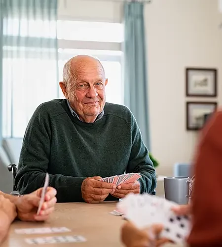 Ein älterer, freundlich lächelnder Mann sitzt an einem Tisch und hält Spielkarten in der Hand; im Vordergrund sind die Hände anderer Mitspieler beim Kartenspiel zu sehen.