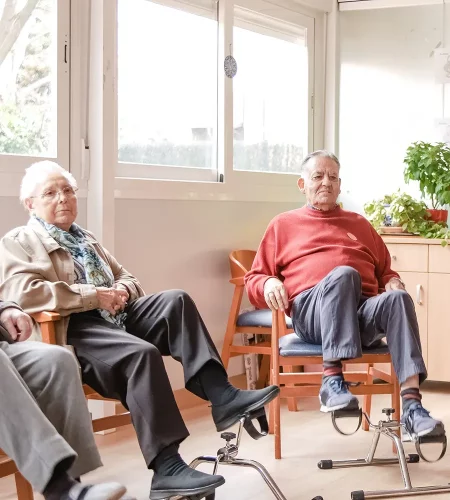 Zwei Senioren sitzen in einem hellen Gemeinschaftsraum auf Stühlen und nutzen Pedaltrainer für ein leichtes Beintraining.