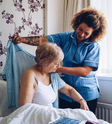 Eine Pflegekraft in blauer Uniform hilft einer älteren Frau im Bett einfühlsam beim Anziehen einer hellblauen Bluse.