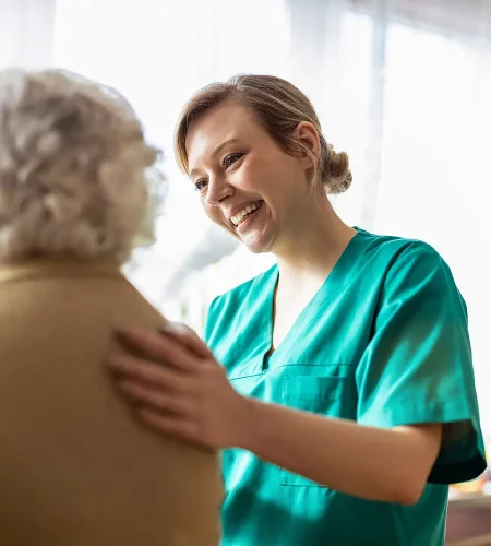 Nahaufnahme einer lächelnden Pflegekraft in grüner Arbeitskleidung, die einer älteren Person im Anschnitt beruhigend die Hand auf die Schulter legt.