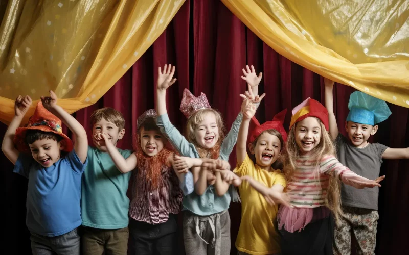 Sieben Kinder tragen bunte Hüte und Umhänge und werfen vor Freude die Arme in die Luft. Sie stehen auf einer kleinen Bühne unter gelben Stoffbahnen direkt vor einem schweren roten Vorhang.